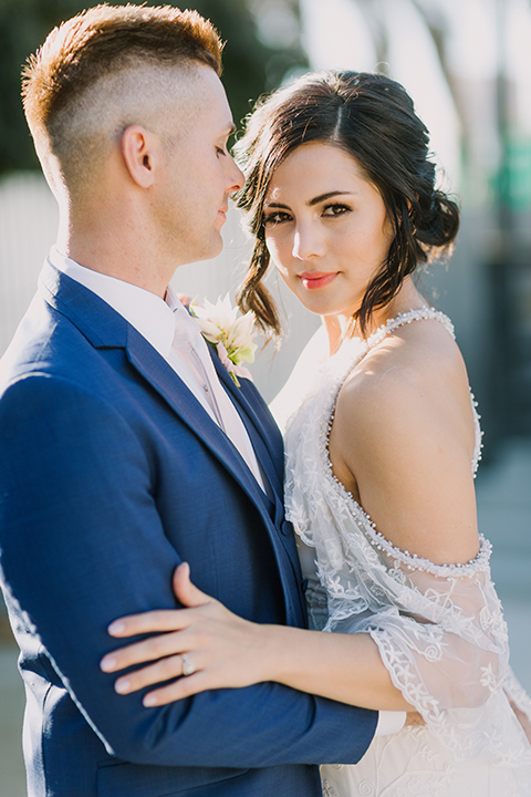 Los angeles outdoor colorful wedding bride a line gown with a high neckline and open shoulder lace sleeves and groom cobalt blue notch lapel suit by allure men with a matching vest and white dress shirt with a long white matte tie and white floral boutonniere standing by table hugging close up