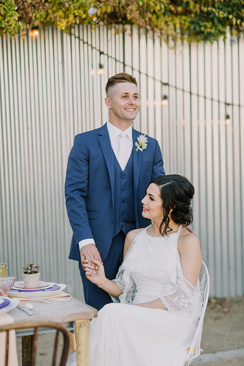 Los angeles outdoor colorful wedding bride a line gown with a high neckline and open shoulder lace sleeves and groom cobalt blue notch lapel suit by allure men with a matching vest and white dress shirt with a long white matte tie and white floral boutonniere standing by table holding hands bride sitting in chair