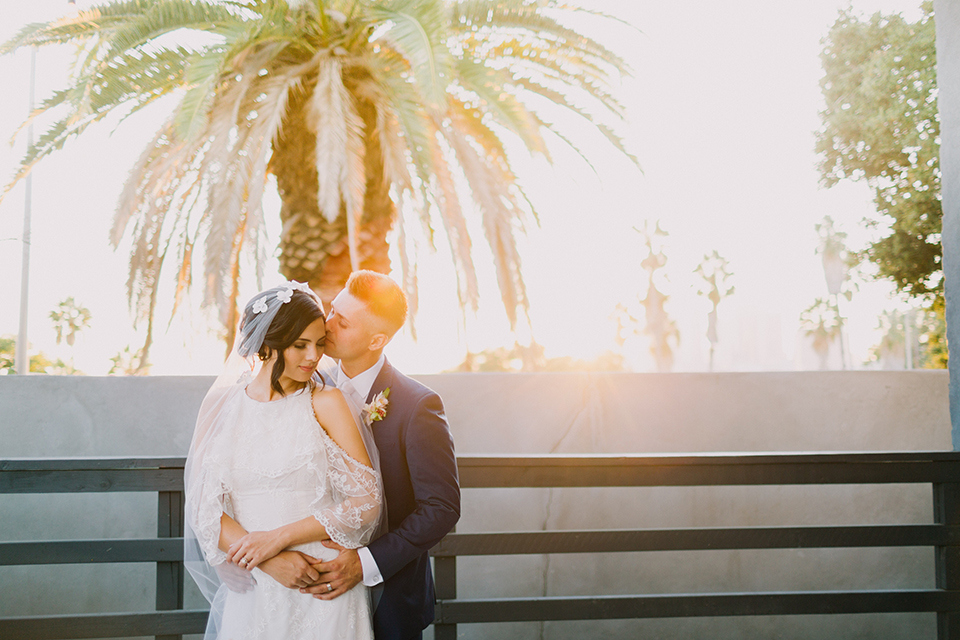 Los angeles outdoor colorful wedding bride a line gown with a high neckline and open shoulder lace sleeves and groom cobalt blue notch lapel suit by allure men with a matching vest and white dress shirt with a long white matte tie and white floral boutonniere standing and hugging sunset