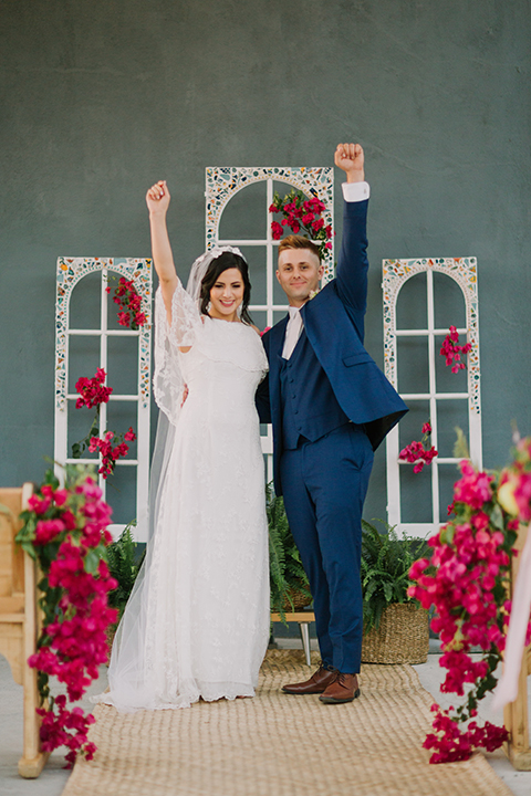 Los angeles outdoor colorful wedding bride a line gown with a high neckline and open shoulder lace sleeves and groom cobalt blue notch lapel suit by allure men with a matching vest and white dress shirt with a long white matte tie and white floral boutonniere cheering after ceremony