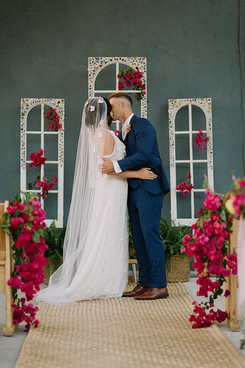 Los angeles outdoor colorful wedding bride a line gown with a high neckline and open shoulder lace sleeves and groom cobalt blue notch lapel suit by allure men with a matching vest and white dress shirt with a long white matte tie and white floral boutonniere kissing during ceremony