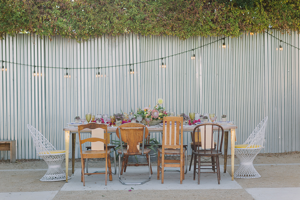 Los angeles outdoor colorful wedding at city libre table set up light brown wood table with an assortment of different style wooden chairs and flower centerpiece decor with white and blue place settings and colorful glassware