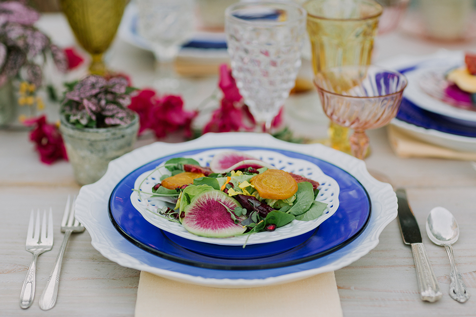 Los angeles outdoor colorful wedding at city libre table set up light brown wood table with an assortment of different style wooden chairs and flower centerpiece decor with white and blue place settings and colorful glassware