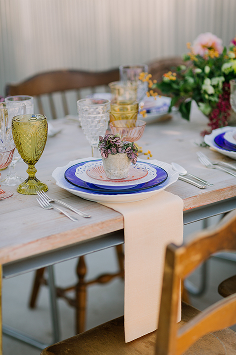 Los angeles outdoor colorful wedding at city libre table set up light brown wood table with an assortment of different style wooden chairs and flower centerpiece decor with white and blue place settings and colorful glassware