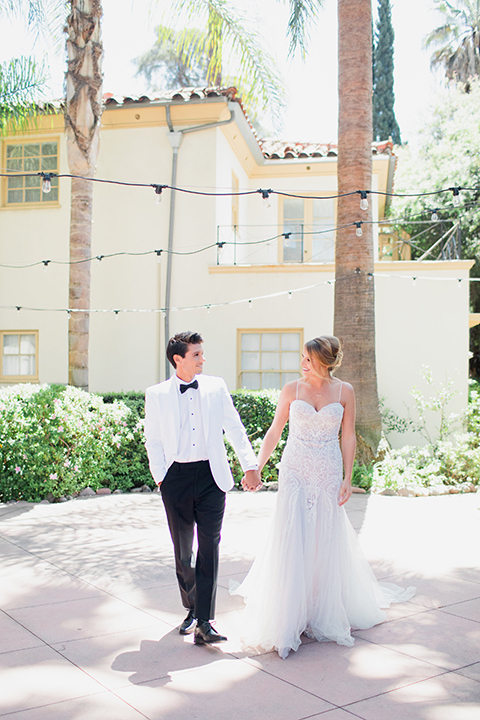 Pasadena outdoor wedding at the maxwell house bride mermaid style gown with thin spaghetti straps and beaded detail with a sweetheart neckline and groom white dinner jacket with black tuxedo pants and a white dress shirt with a black bow tie holding hands and walking