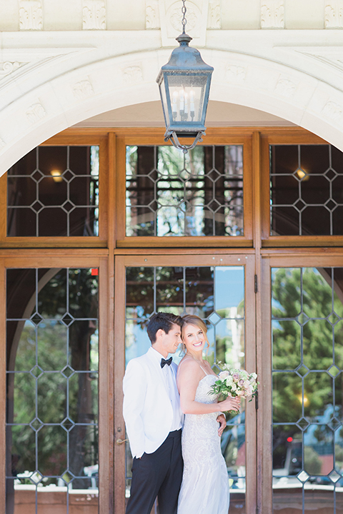 Pasadena outdoor wedding at the maxwell house bride mermaid style gown with thin spaghetti straps and beaded detail with a sweetheart neckline and groom white dinner jacket with black tuxedo pants and a white dress shirt with a black bow tie hugging and bride holding white and green floral bridal bouquet