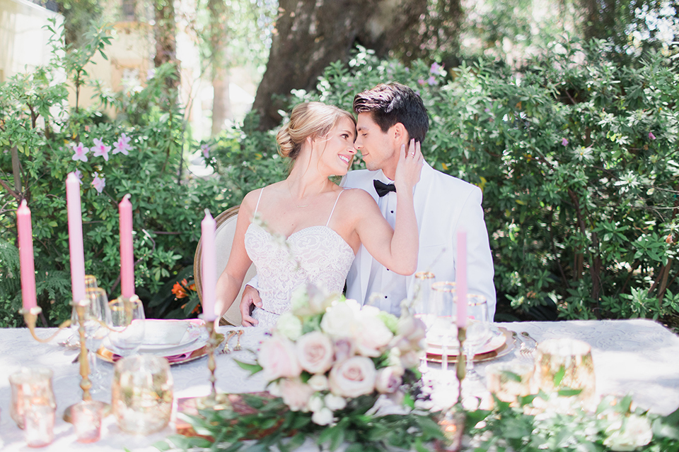 Pasadena outdoor wedding at the maxwell house bride mermaid style gown with thin spaghetti straps and beaded detail with a sweetheart neckline and groom white dinner jacket with black tuxedo pants and a white dress shirt with a black bow tie sitting by table hugging