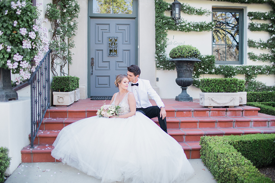 Pasadena outdoor wedding at the maxwell house bride mermaid style gown with thin spaghetti straps and beaded detail with a sweetheart neckline and groom white dinner jacket with black tuxedo pants and a white dress shirt with a black bow tie sitting on steps smiling