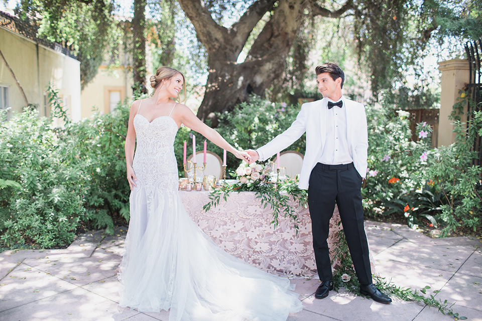 Pasadena outdoor wedding at the maxwell house bride mermaid style gown with thin spaghetti straps and beaded detail with a sweetheart neckline and groom white dinner jacket with black tuxedo pants and a white dress shirt with a black bow tie standing by table holding hands