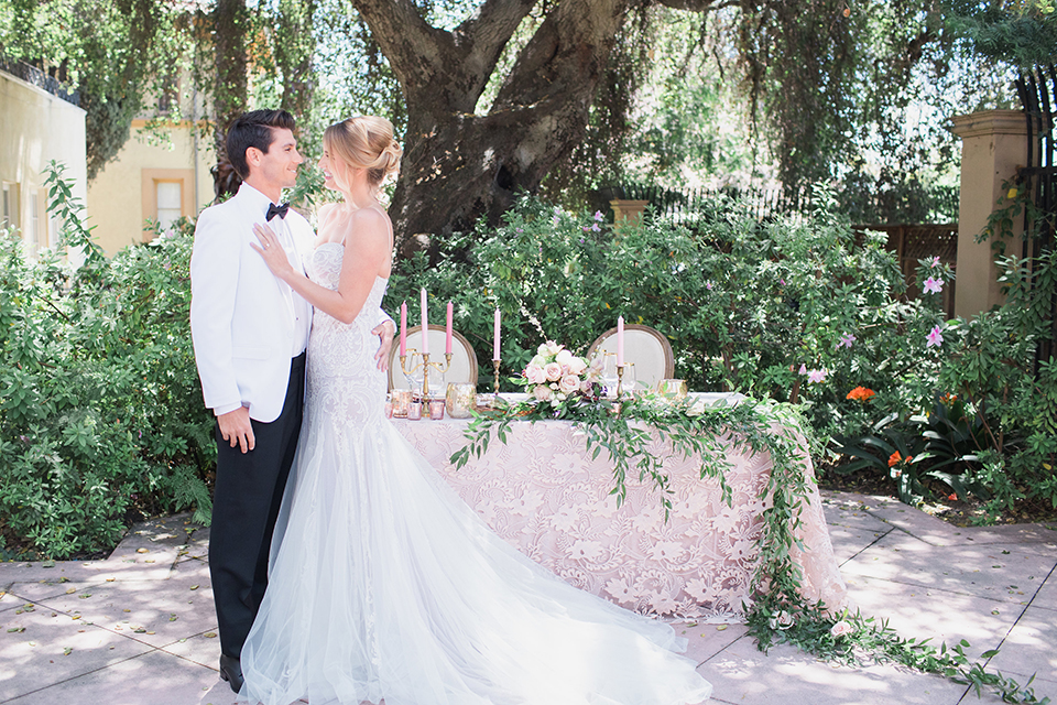 Pasadena outdoor wedding at the maxwell house bride mermaid style gown with thin spaghetti straps and beaded detail with a sweetheart neckline and groom white dinner jacket with black tuxedo pants and a white dress shirt with a black bow tie standing by table hugging