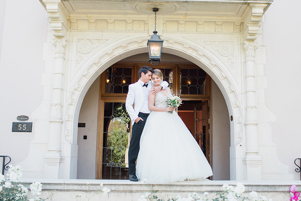 Pasadena outdoor wedding at the maxwell house bride mermaid style gown with thin spaghetti straps and beaded detail with a sweetheart neckline and groom white dinner jacket with black tuxedo pants and a white dress shirt with a black bow tie standing on ledge hugging