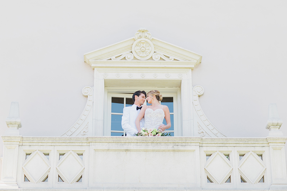 Pasadena outdoor wedding at the maxwell house bride mermaid style gown with thin spaghetti straps and beaded detail with a sweetheart neckline and groom white dinner jacket with black tuxedo pants and a white dress shirt with a black bow tie standing on balcony hugging