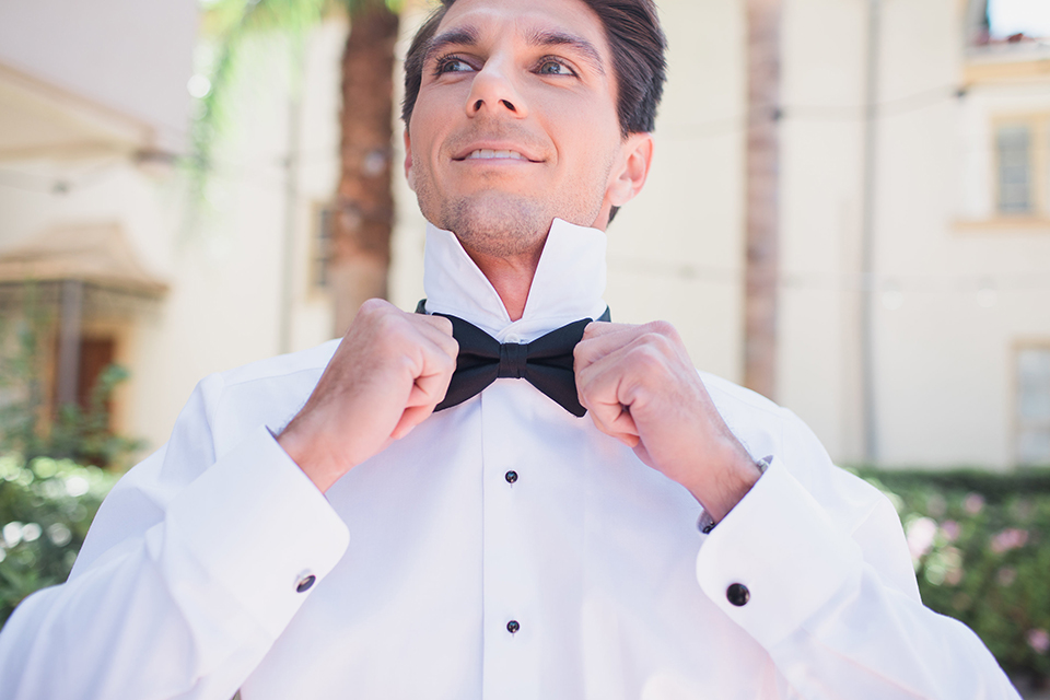 Pasadena outdoor wedding at the maxwell house groom white dinner jacket with black tuxedo pants and a white dress shirt with a black bow tie getting ready holding bow tie