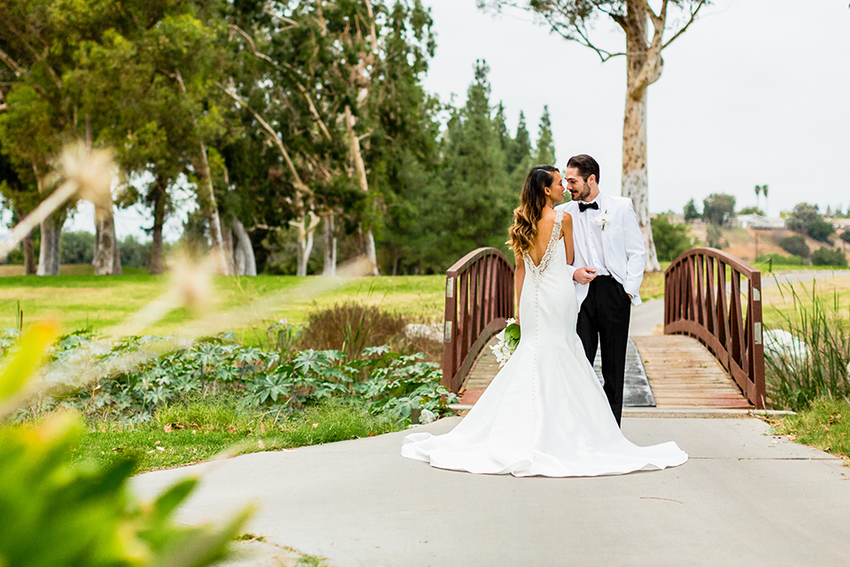 Southern california outdoor wedding at diamond bar golf course bride form fitting mermaid style gown with silver beaded straps and a sweetheart neckline with a low back design and beaded detail with groom white notch lapel dinner jacket with a white dress shirt and black tuxedo pants with a black bow tie hugging