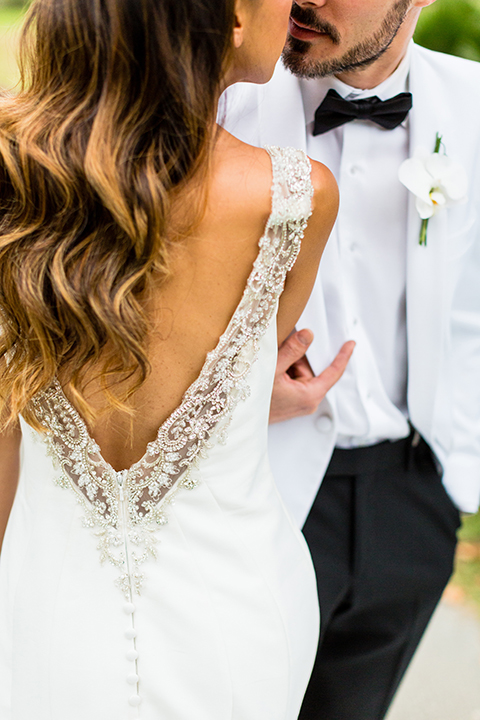 Southern california outdoor wedding at diamond bar golf course bride form fitting mermaid style gown with silver beaded straps and a sweetheart neckline with a low back design and beaded detail with groom white notch lapel dinner jacket with a white dress shirt and black tuxedo pants with a black bow tie hugging and holding hands close up