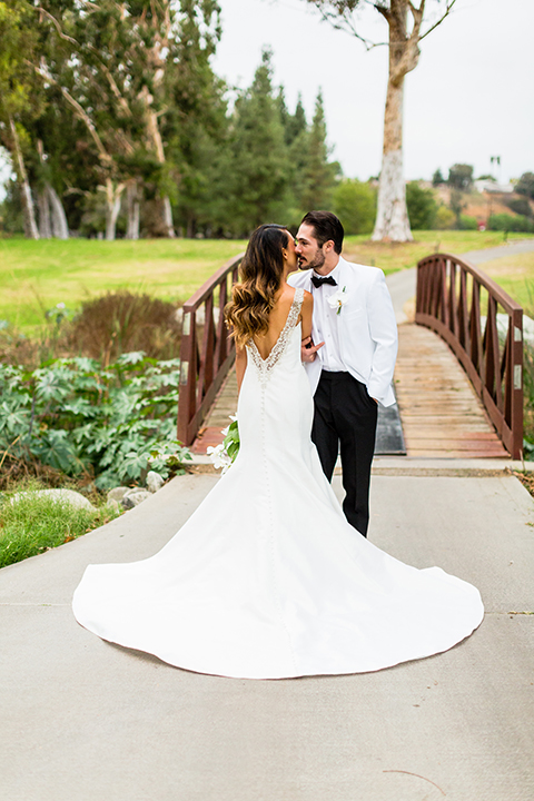 Southern california outdoor wedding at diamond bar golf course bride form fitting mermaid style gown with silver beaded straps and a sweetheart neckline with a low back design and beaded detail with groom white notch lapel dinner jacket with a white dress shirt and black tuxedo pants with a black bow tie hugging 