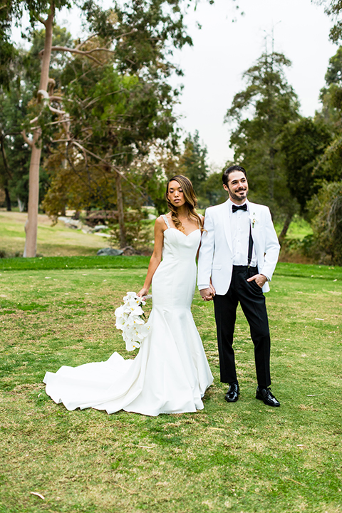 Southern california outdoor wedding at diamond bar golf course bride form fitting mermaid style gown with silver beaded straps and a sweetheart neckline with a low back design and beaded detail with groom white notch lapel dinner jacket with a white dress shirt and black tuxedo pants with a black bow tie hugging and bride holding white and green floral bridal bouquet