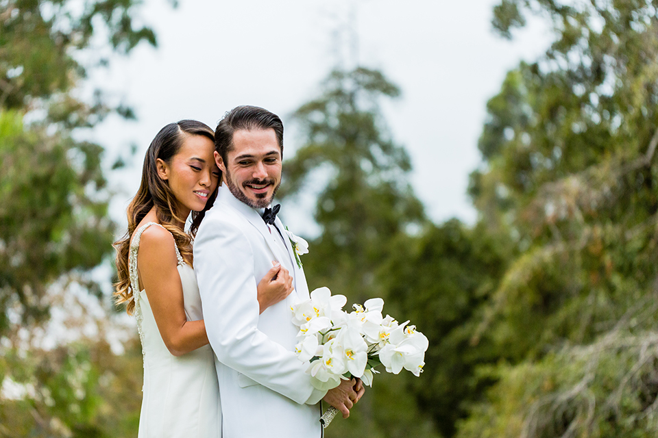 Southern california outdoor wedding at diamond bar golf course bride form fitting mermaid style gown with silver beaded straps and a sweetheart neckline with a low back design and beaded detail with groom white notch lapel dinner jacket with a white dress shirt and black tuxedo pants with a black bow tie hugging groom holding white and green floral bridal bouquet