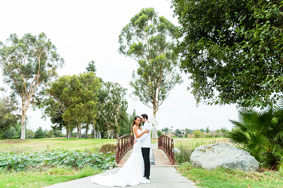 Southern california outdoor wedding at diamond bar golf course bride form fitting mermaid style gown with silver beaded straps and a sweetheart neckline with a low back design and beaded detail with groom white notch lapel dinner jacket with a white dress shirt and black tuxedo pants with a black bow tie hugging far away