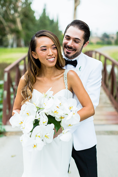 Southern california outdoor wedding at diamond bar golf course bride form fitting mermaid style gown with silver beaded straps and a sweetheart neckline with a low back design and beaded detail with groom white notch lapel dinner jacket with a white dress shirt and black tuxedo pants with a black bow tie hugging close up and bride holding white and green floral bridal bouquet