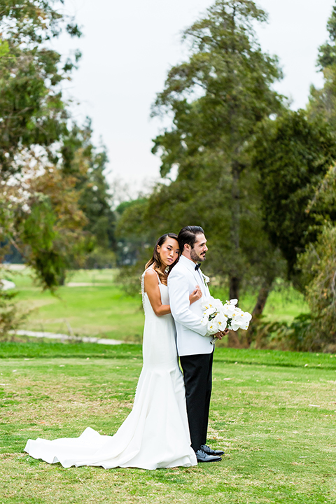 Southern california outdoor wedding at diamond bar golf course bride form fitting mermaid style gown with silver beaded straps and a sweetheart neckline with a low back design and beaded detail with groom white notch lapel dinner jacket with a white dress shirt and black tuxedo pants with a black bow tie bride hugging groom behind and groom holding white and green floral bridal bouquet