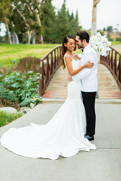 Southern california outdoor wedding at diamond bar golf course bride form fitting mermaid style gown with silver beaded straps and a sweetheart neckline with a low back design and beaded detail with groom white notch lapel dinner jacket with a white dress shirt and black tuxedo pants with a black bow tie hugging and bride holding white and green floral bridal bouquet