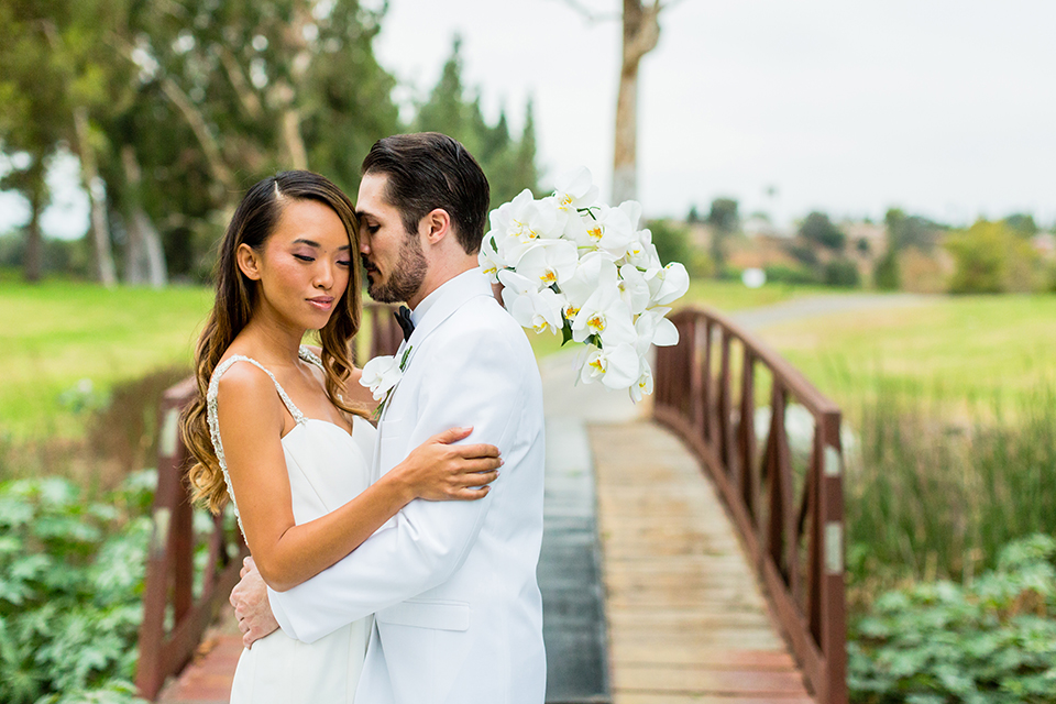 Southern california outdoor wedding at diamond bar golf course bride form fitting mermaid style gown with silver beaded straps and a sweetheart neckline with a low back design and beaded detail with groom white notch lapel dinner jacket with a white dress shirt and black tuxedo pants with a black bow tie hugging and bride holding white and green floral bridal bouquet