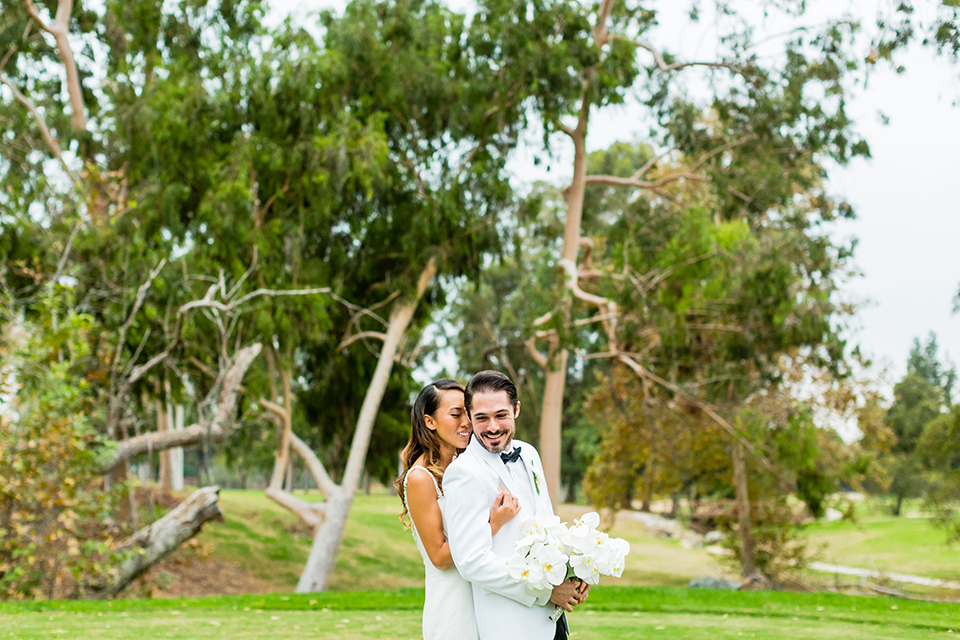 Southern california outdoor wedding at diamond bar golf course bride form fitting mermaid style gown with silver beaded straps and a sweetheart neckline with a low back design and beaded detail with groom white notch lapel dinner jacket with a white dress shirt and black tuxedo pants with a black bow tie bride hugging groom and groom holding white and green floral bridal bouquet