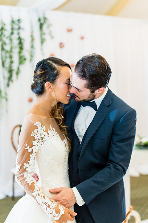 Southern california outdoor wedding at diamond bar golf course bride ball gown style dress with lace illusion long sleeves and illusion back with buttons with groom slate blue notch lapel suit by allure men with a matching vest and white dress shirt and matching blue bow tie standing by table kissing