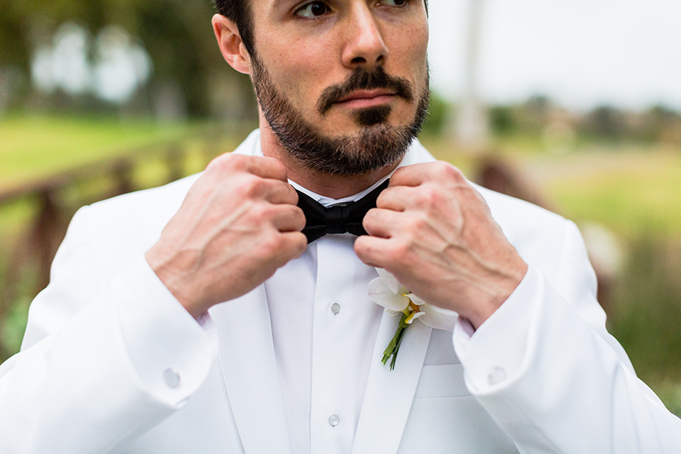 Southern california outdoor wedding at diamond bar golf course groom white notch lapel dinner jacket with a white dress shirt and black tuxedo pants with a black bow tie and white floral boutonniere holding bow tie