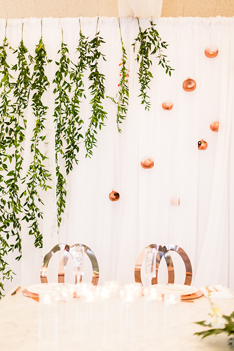 Southern california outdoor wedding at diamond bar golf course table set up white table with greenery floral decor and rose gold chairs with hanging decor and wall with greenery florals and white and gold place settings with gold silverware