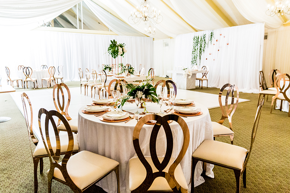 Southern california outdoor wedding at diamond bar golf course table set up white table with greenery floral decor and rose gold chairs with hanging decor and wall with greenery florals and white and gold place settings with gold silverware