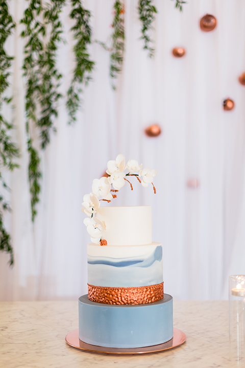 Southern california outdoor wedding at diamond bar golf course table set up white table with greenery floral decor and rose gold chairs with hanging decor and wall with greenery florals and white and gold place settings with gold silverware three tier wedding cake with blue and white designs