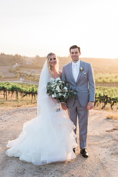 Temecula outdoor wedding at falkner winery bride mermaid style gown with lace bodice and sweetheart neckline with ruffled skirt and long veil holding white and green floral bridal bouquet in vineyard