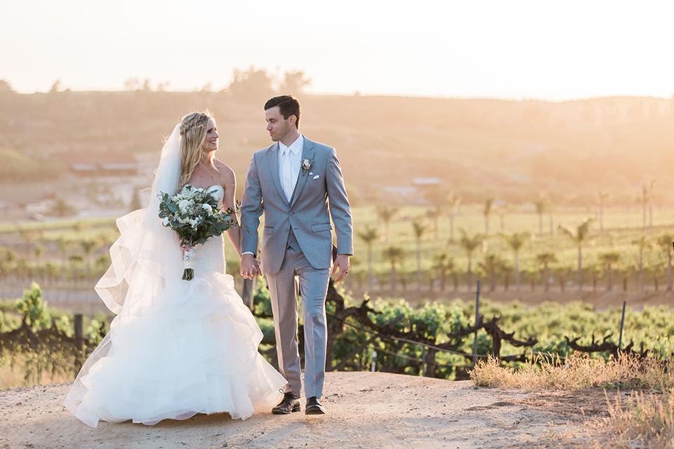 Temecula outdoor wedding at falkner winery bride mermaid style gown with lace bodice and sweetheart neckline with ruffled skirt and long veil holding white and green floral bridal bouquet in vineyard holding hands and walking