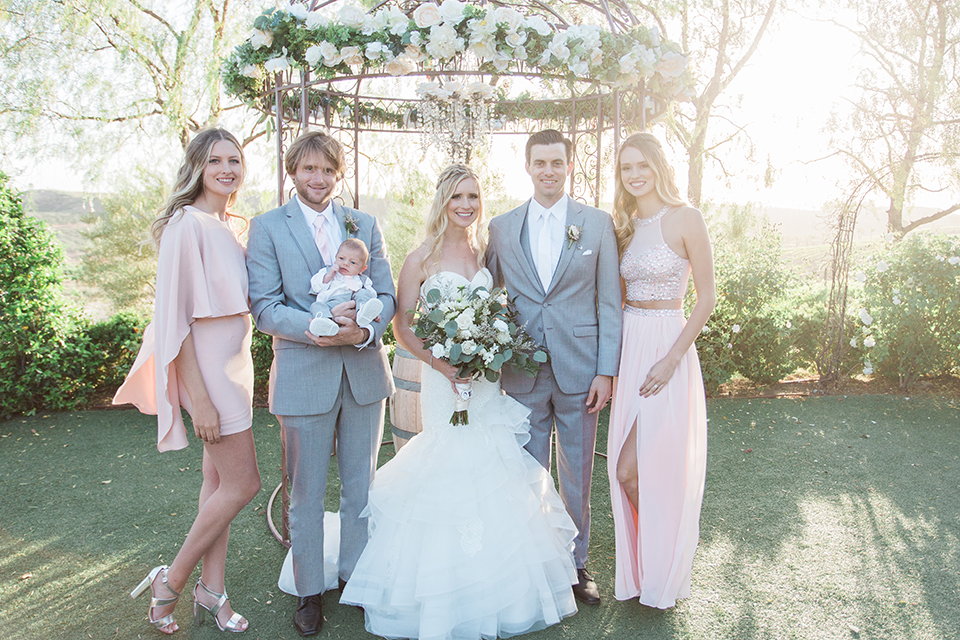 Temecula outdoor wedding at falkner winery bride mermaid style gown with lace bodice and sweetheart neckline with ruffled skirt and long veil holding white and green floral bridal bouquet standing with family smiling