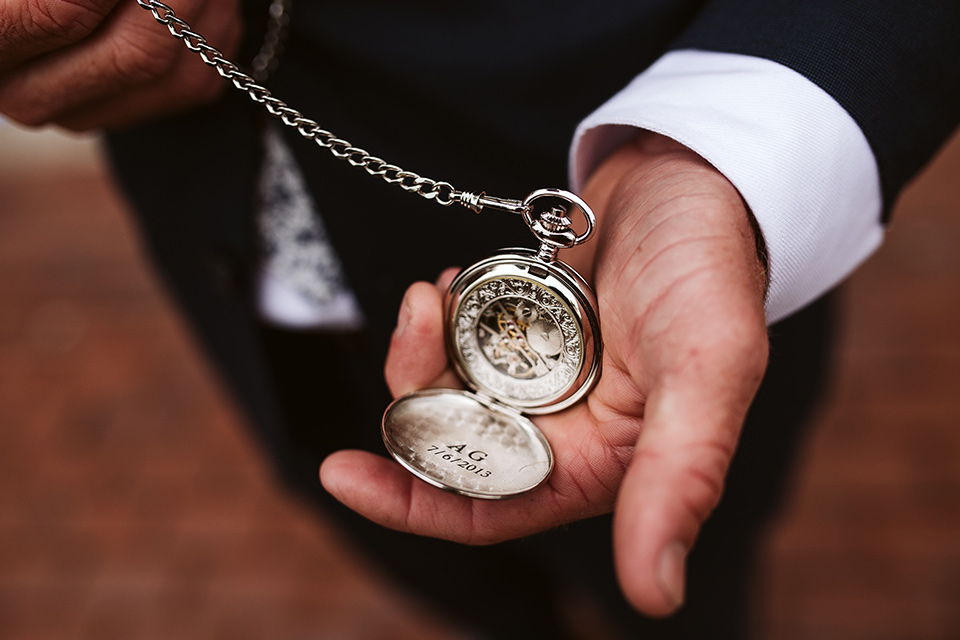 Adobe-de-Capastrano-Amanda-and-Asher-pocketwatch-groom-wearing-a-navy-suit-with-floral-tie-holding-a-pocketwatch-that-is-engraved-with-their-wedding-date