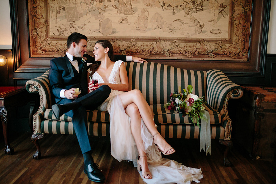 Southern-california-wedding-at-the-ebell-of-long-beach-bride-and-groom-sitting-on-couch-bride-wearing-a-lace-a-line-dress-with-straps-and-hair-in-loose-down-waves-and-groom-in-navy-tux-with-black-trim-and-black-bowtie