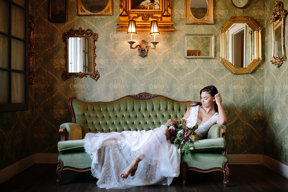 Southern-california-wedding-at-the-ebell-of-long-beach-bride-lace-gown-sitting-bride-wearing-a-lace-a-line-dress-with-straps-and-hair-in-loose-down-waves-holding-her-bouquet-and-her-hand-resting-on-her-head