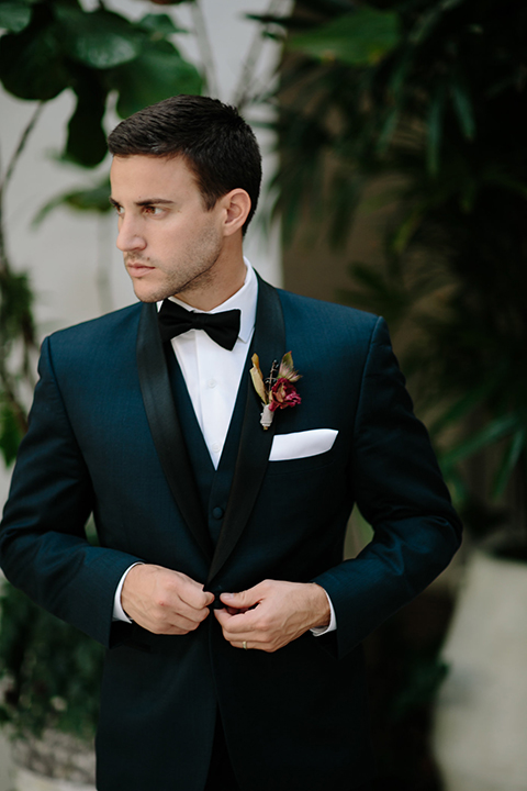 Southern-california-wedding-at-the-ebell-of-long-beach-groom-navy-tuxedo-buttoning-jacket-groom-in-navy-tux-with-black-trim-and-black-bowtie-with-a-white-pocket-square-and-burgundy-boutionniere