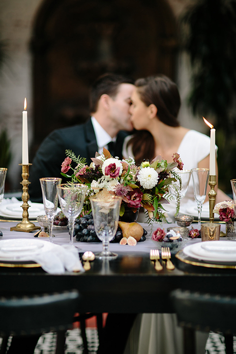 Southern-california-wedding-at-the-ebell-of-long-beach-reception-bride-and-groom-sitting-at-table-kissing-bride-wearing-a-lace-a-line-dress-with-straps-and-hair-in-loose-down-waves-and-groom-in-navy-tux-with-black-trim-and-black-bowtie