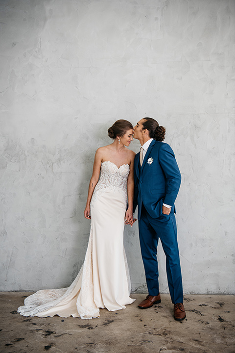 FD-Studios-bride-and-groom-kissing-by-grey-wall-bride-wearing-a-flowing-strapless-gown-with-a-lace-bodice-groom-in-a-dark-blue-suit-with-a-blush-long-tie