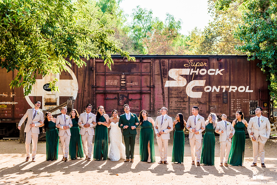 Real-wedding-lani-and-ryan-all-the-bridal-party-arms-linked-and-laughing-groomsmen-in-tan-suits-with-floral-ties-and-brown-shoes-groom-in-green-suit-with-a-floral-tie-and-brown-shoes-bridesmaids-are-in-dark-green-dresses-with-lace-detailing-and-halter-nekline-bride-in-a-lace-a-line-dress-with-hair-in-a-bun-and-long-veil