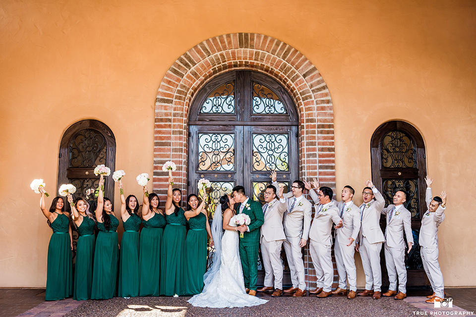 Real-wedding-lani-and-ryan-bridal-party-by-terrecotta-wall-groomsmen-in-tan-suits-with-floral-ties-and-brown-shoes-groom-in-green-suit-with-a-floral-tie-and-brown-shoes-bridesmaids-are-in-dark-green-dresses-with-lace-detailing-and-halter-nekline-bride-in-a-lace-a-line-dress-with-hair-in-a-bun-and-long-veil