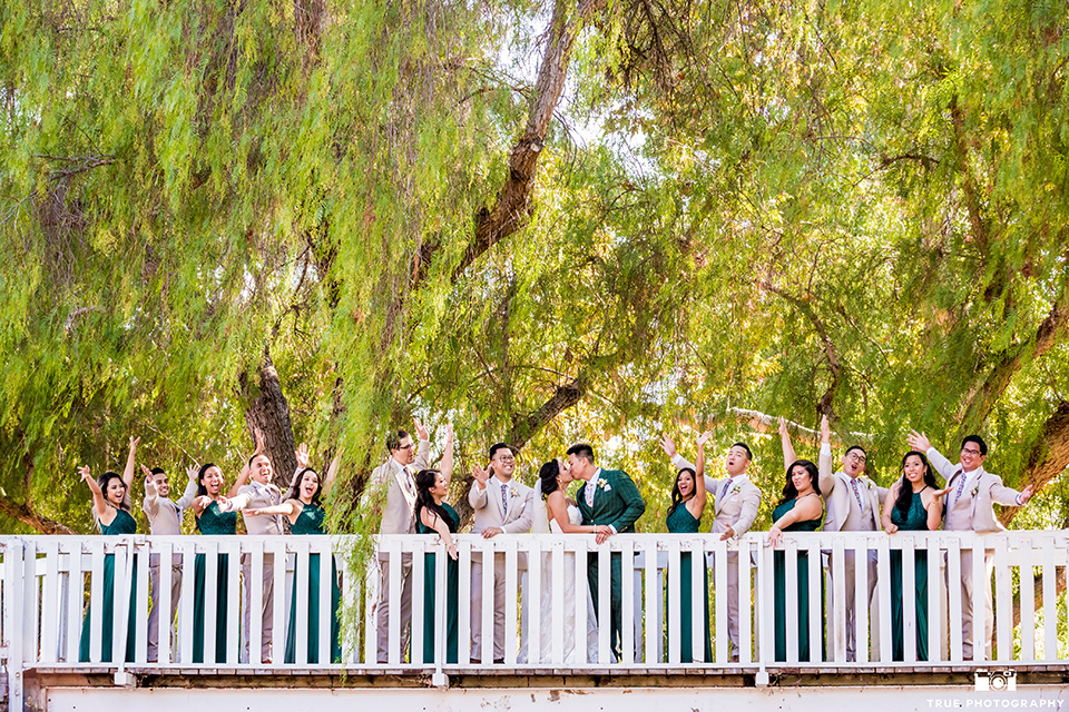 Real-wedding-lani-and-ryan-bridal-party-on-white-bridge-bride-and-groom-kissing-groomsmen-in-tan-suits-with-floral-ties-and-brown-shoes-groom-in-green-suit-with-a-floral-tie-and-brown-shoes-bridesmaids-are-in-dark-green-dresses-with-lace-detailing-and-halter-nekline-bride-in-a-lace-a-line-dress-with-hair-in-a-bun-and-long-veil