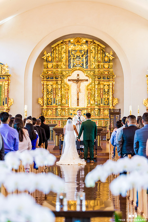Real-wedding-lani-and-ryan-bride-and-groom-holding-hands-at-the-altar-backs-facing-the-camera-groom-in-green-suit-with-a-floral-tie-and-brown-shoes-bride-in-a-lace-a-line-dress-with-hair-in-a-bun-and-long-veil