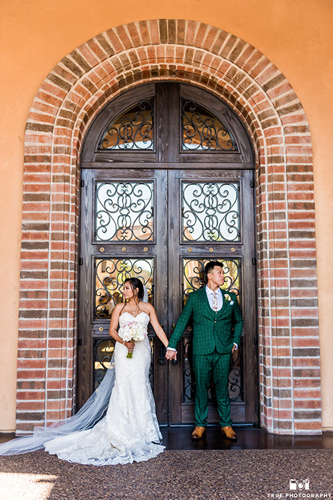Real-wedding-lani-and-ryan-bride-and-groom-holding-hands-facing-away-from-each-other-groom-in-green-suit-with-a-floral-tie-and-brown-shoes-bride-in-a-lace-a-line-dress-with-hair-in-a-bun-and-long-veil