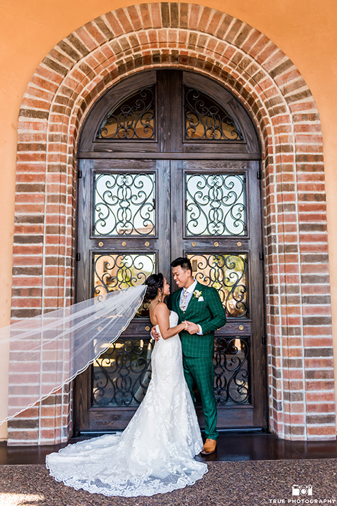 Real-wedding-lani-and-ryan-by-brick-wall-looking-at-each-other-groom-in-green-suit-with-a-floral-tie-and-brown-shoes-bride-in-a-lace-a-line-dress-with-hair-in-a-bun-and-long-veil