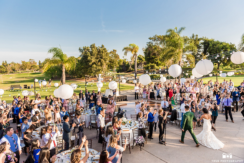 Real-wedding-lani-and-ryan-entering-their-recpetion-overhead-photo-groomsmen-in-tan-suits-with-floral-ties-and-brown-shoes-groom-in-green-suit-with-a-floral-tie-and-brown-shoes-bridesmaids-are-in-dark-green-dresses-with-lace-detailing-and-halter-nekline-bride-in-a-lace-a-line-dress-with-hair-in-a-bun-and-long-veil