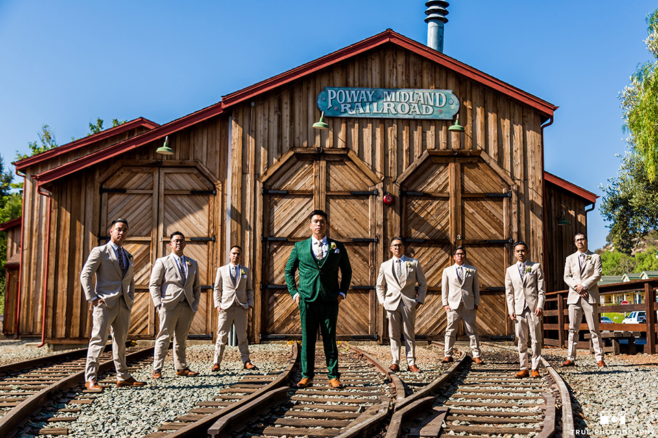 Real-wedding-lani-and-ryan-groomsmen-and-groom-standing-with-hands-in-pocket-in-front-of-train-depot-groomsmen-in-tan-suits-with-floral-ties-and-brown-shoes-groom-in-green-suit-with-a-floral-tie-and-brown-shoes